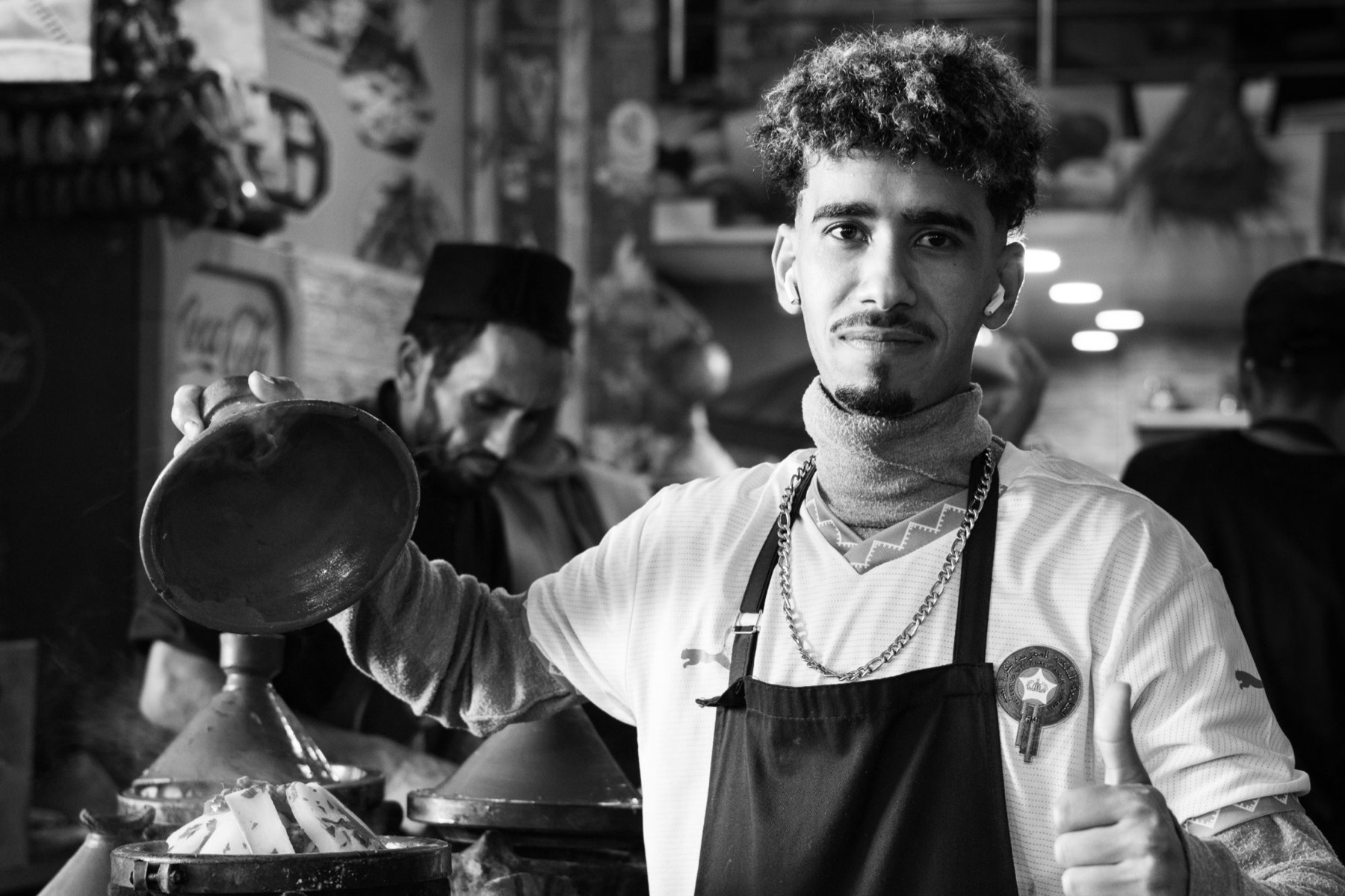 Schwarzweiß: junger Koch mit Schürze hebt den Deckel einer Tajine und zeigt den Daumen nach oben